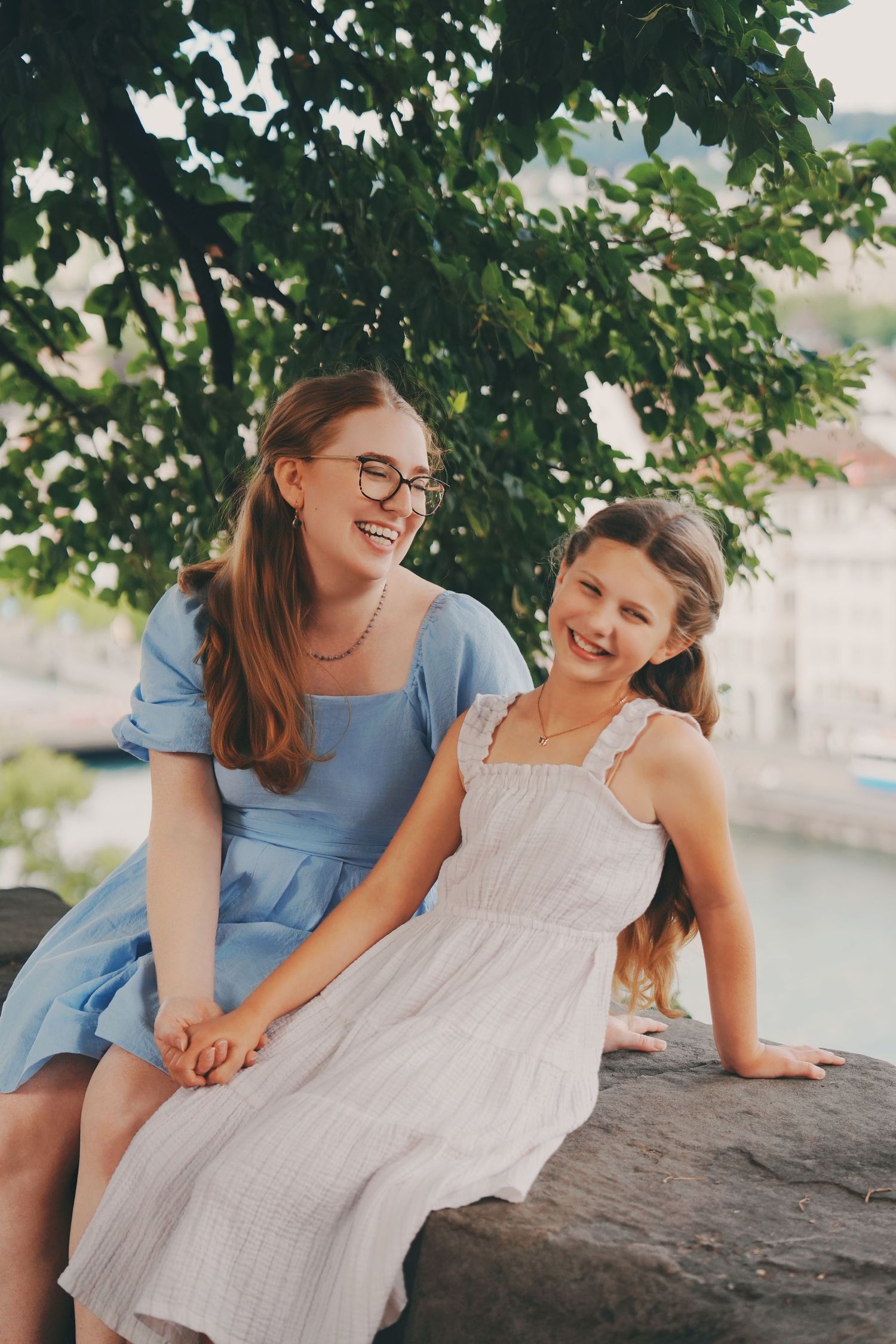Two happy girls in summer dresses sitting on a stone wall laughing together.