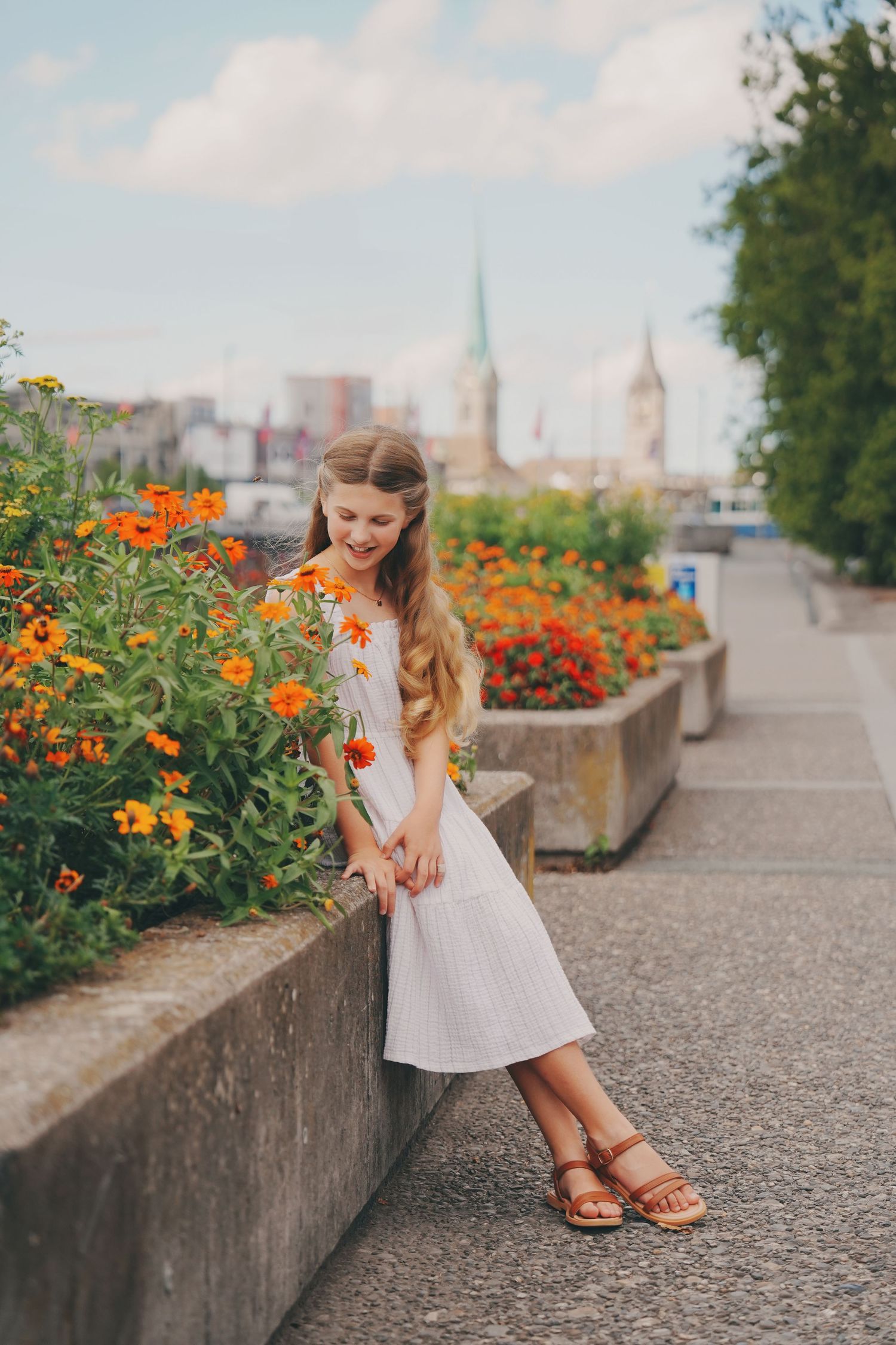 Young girl in white dress standing beside vibrant orange flowers with city skyline behind.
