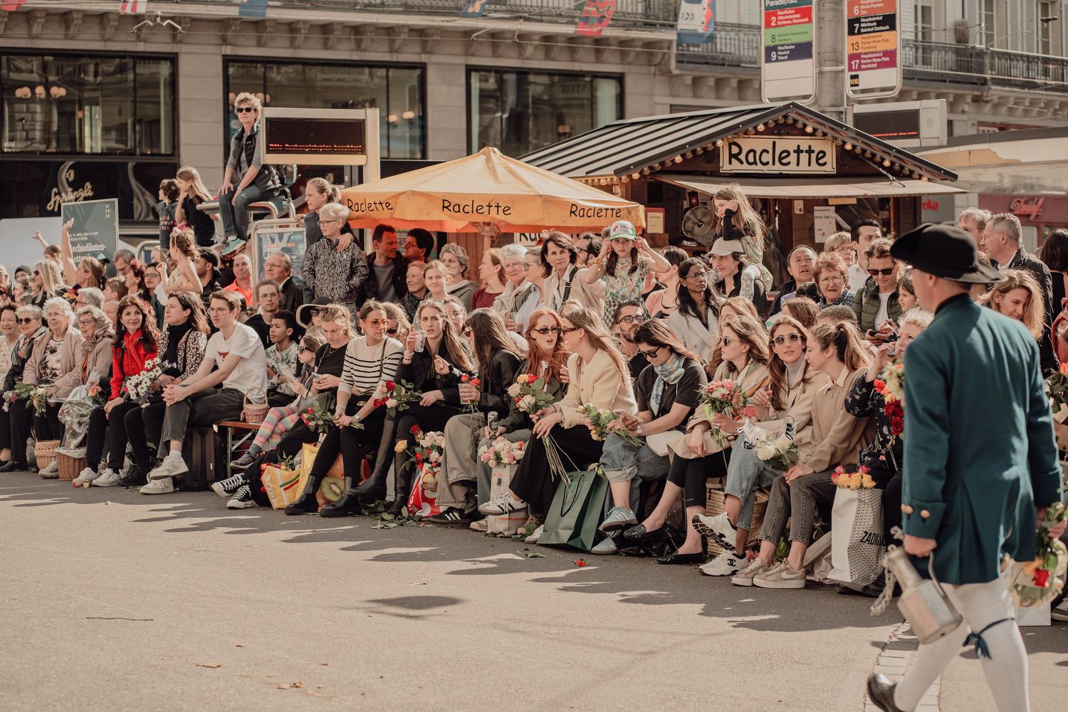 A large crowd of spectators watches a traditional folk parade near a Raclette food stall outdoors.