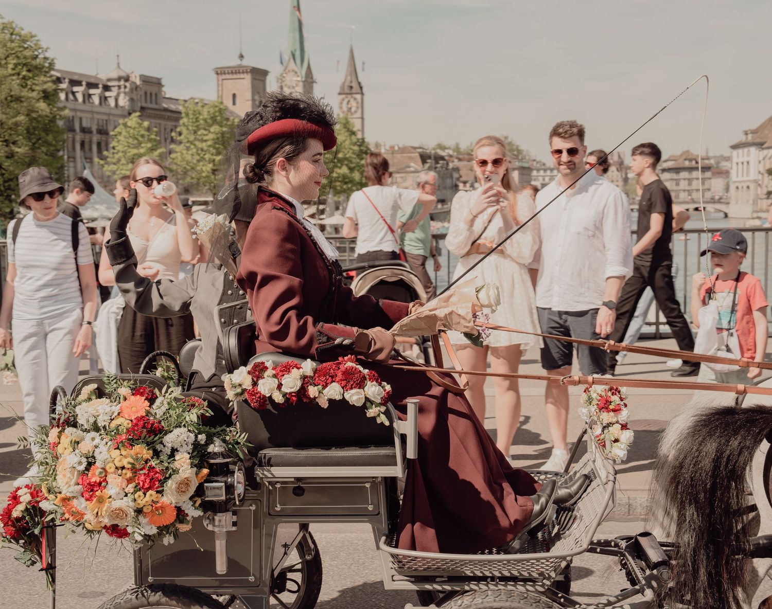 Woman in historical burgundy outfit steering a floral carriage through a crowded Zurich street.