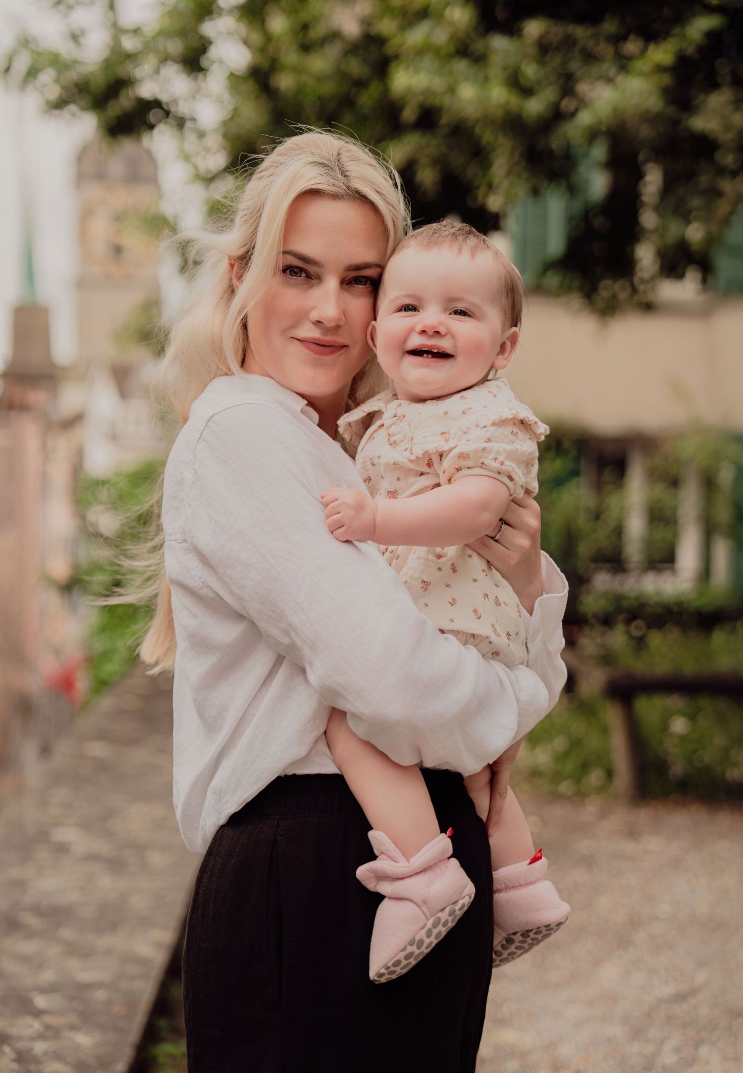 Smiling blonde mother holding laughing baby girl on charming European cobblestone street.