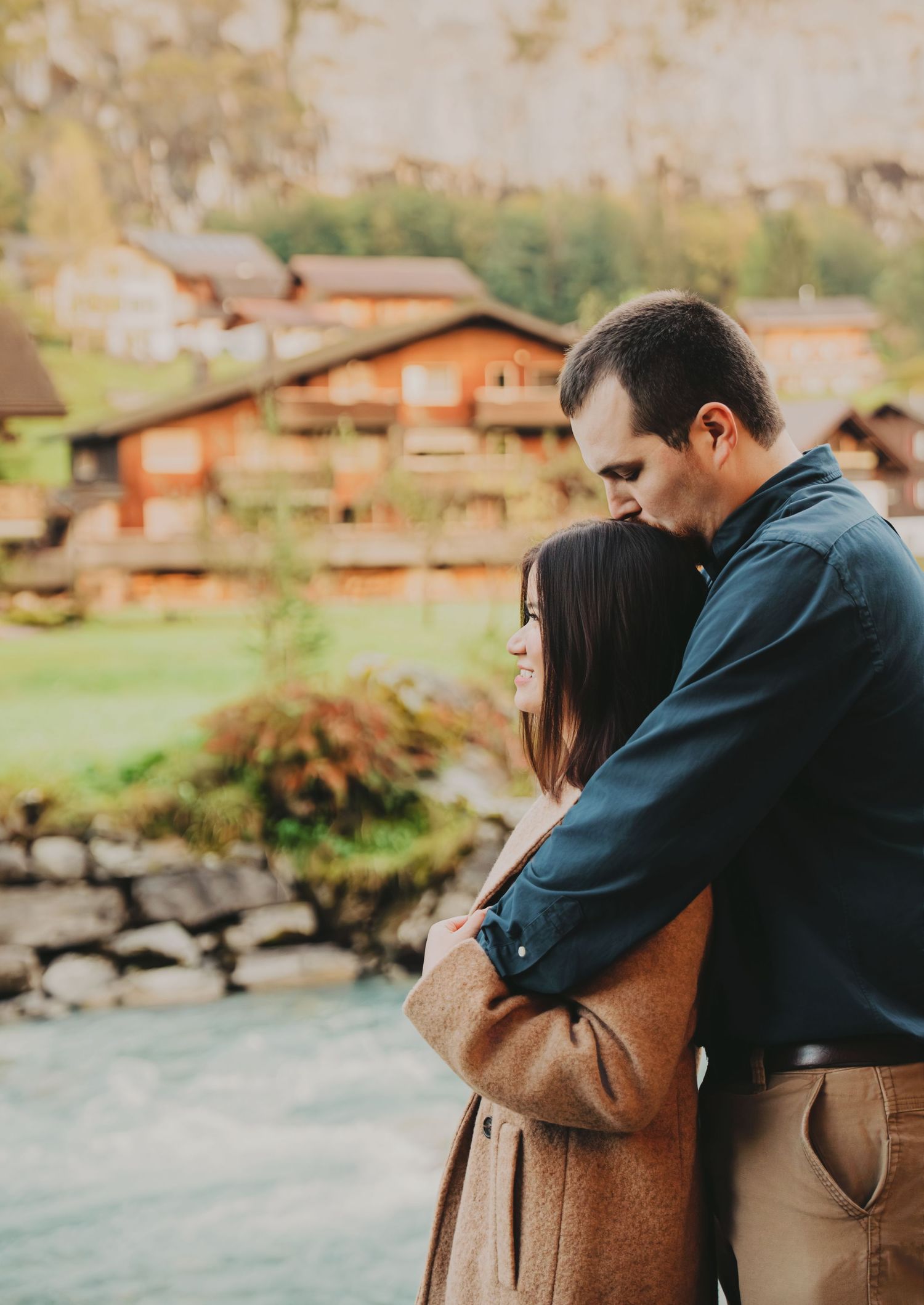 Man tenderly kissing woman's head while embracing near a Swiss alpine river village.