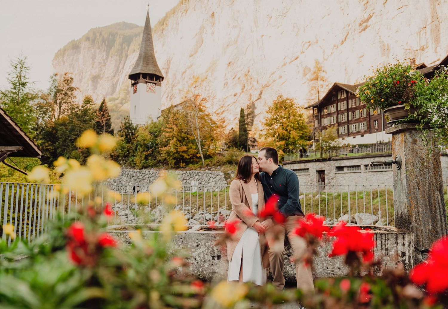 Couple kissing near colorful flowers with a Swiss church steeple and cliff backdrop.