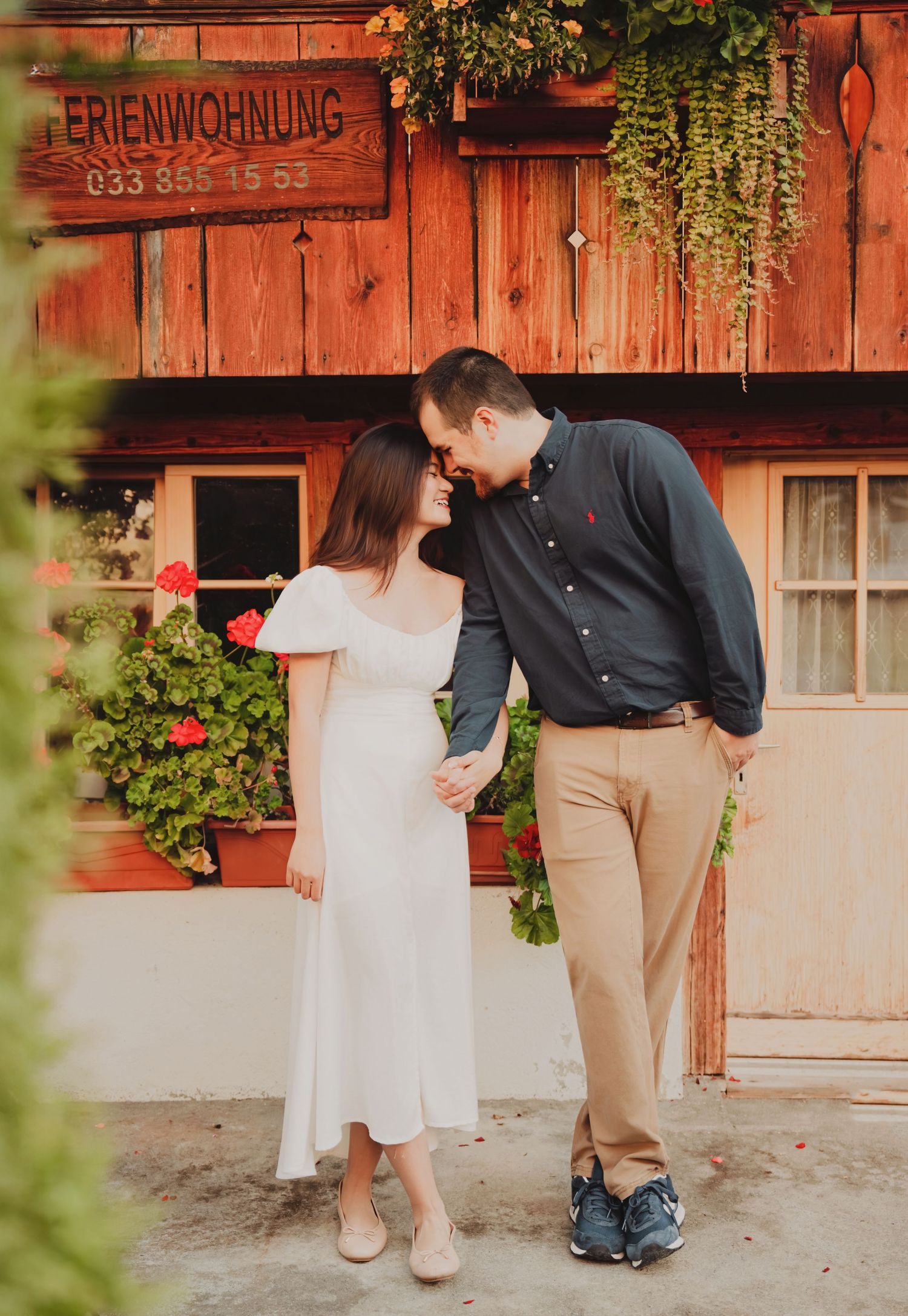Laughing couple holding hands outside a rustic red Swiss Ferienwohnung building with flowers.