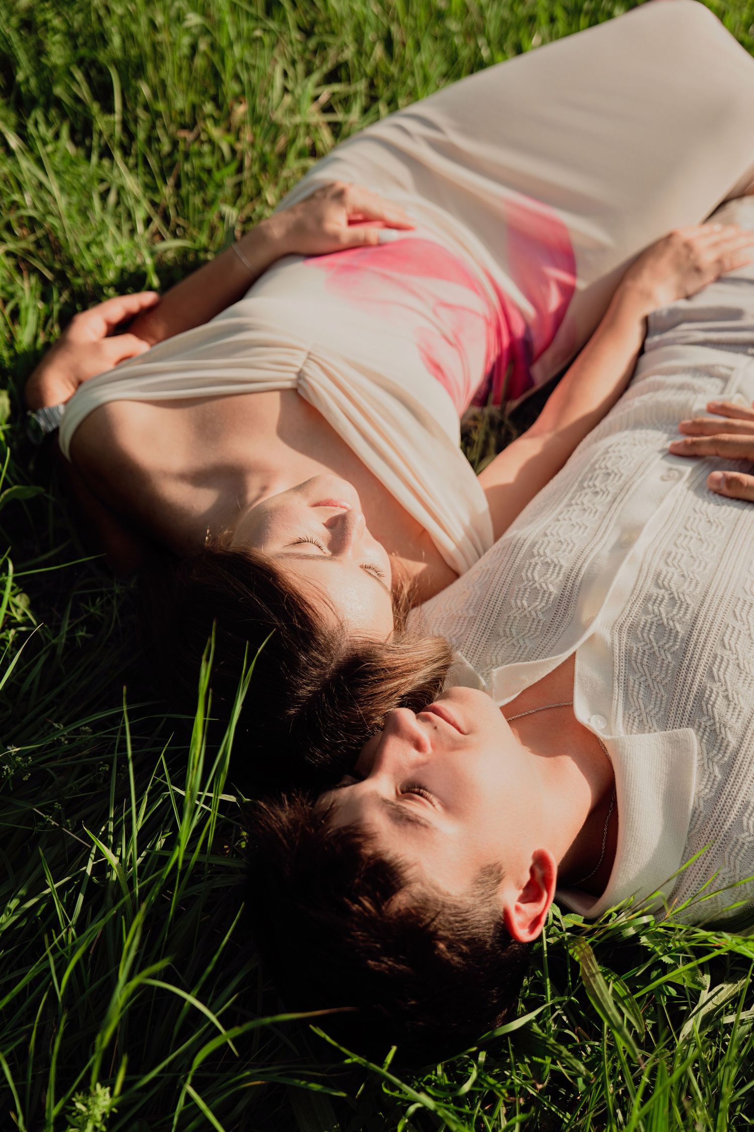 A romantic couple lies together in green grass, basking in warm sunlight outdoors.