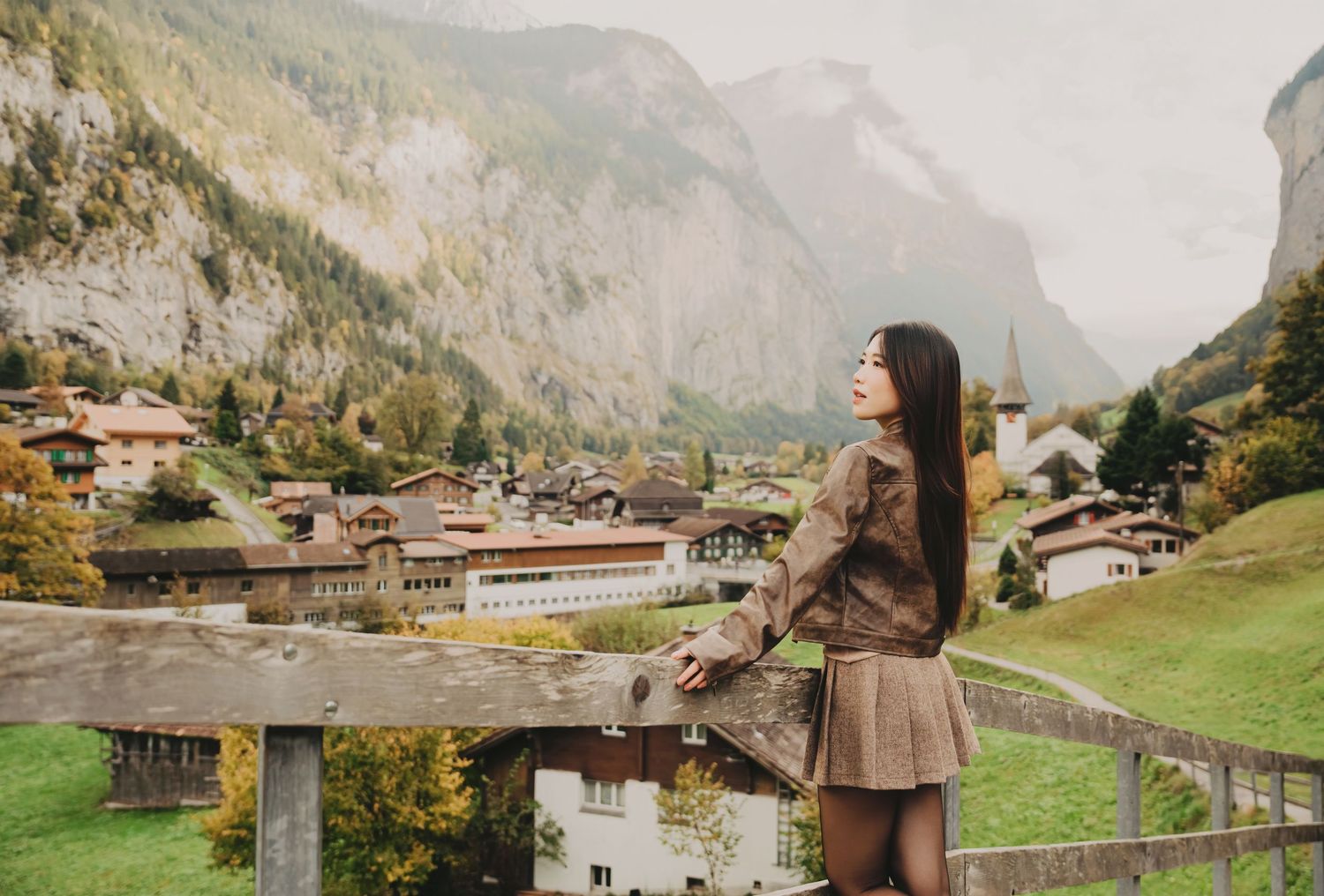 Young woman in brown outfit leaning on a wooden fence with a charming Swiss alpine village behind her.