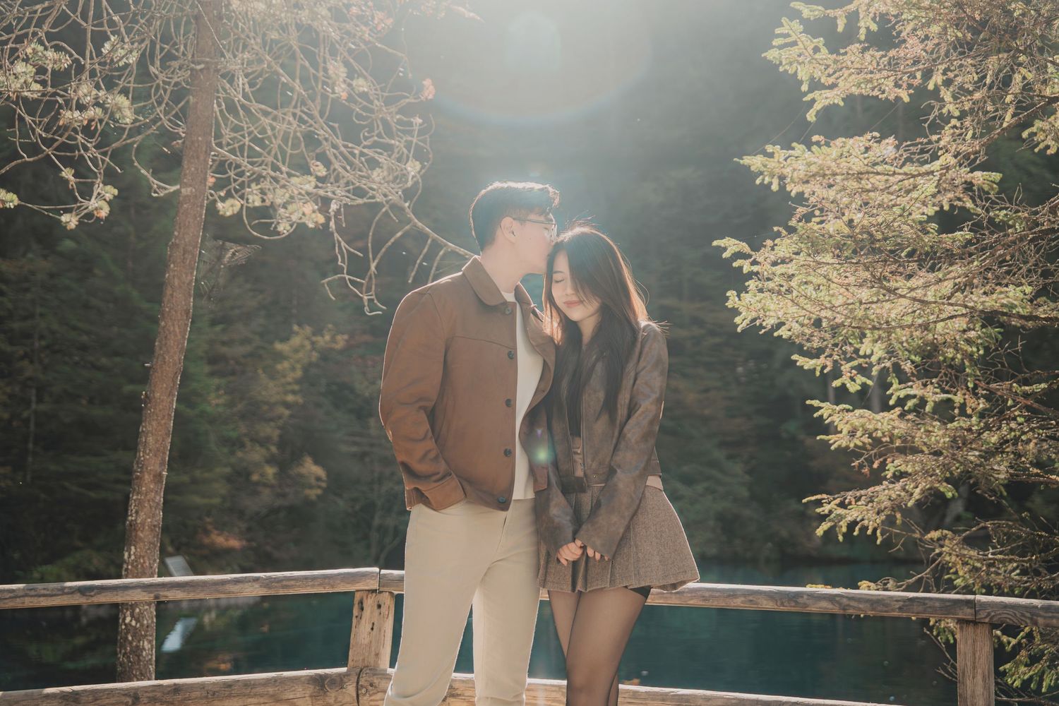 Romantic couple sharing a kiss on a wooden deck surrounded by tall pine trees in sunlight.