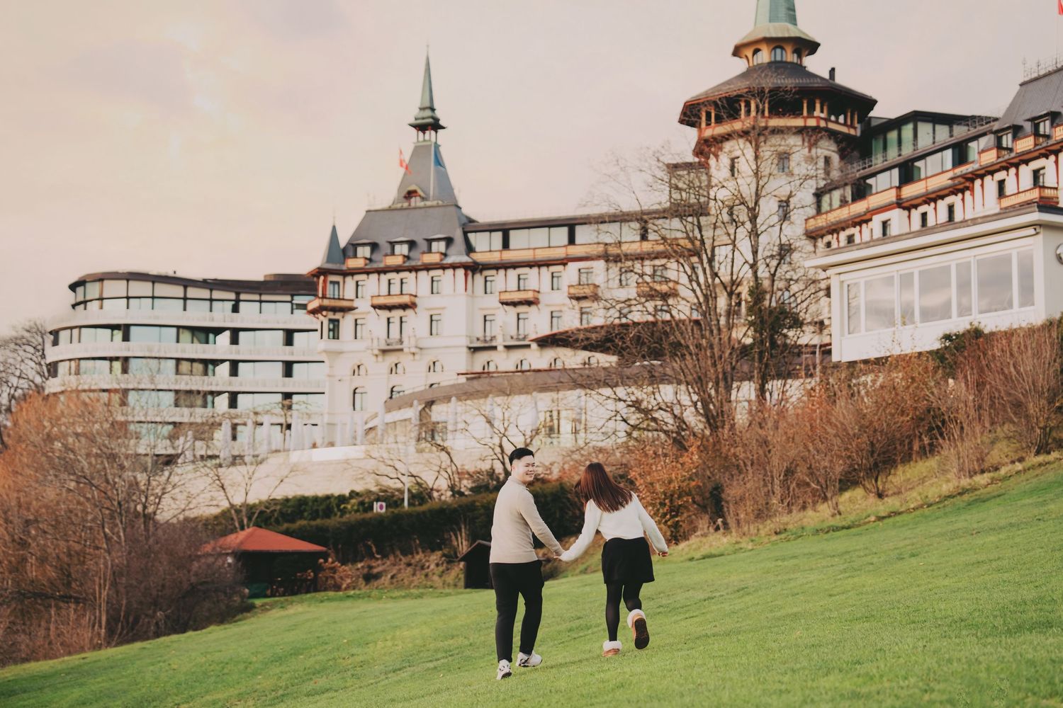 Couple holding hands walking toward grand European castle hotel on hillside.