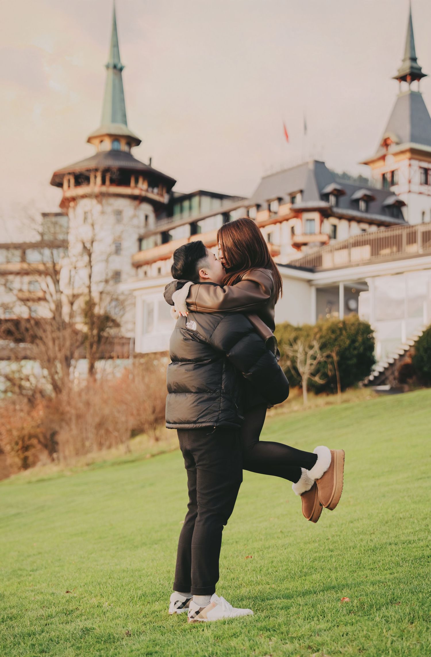 Man lifting woman in romantic embrace on green lawn before grand European castle.