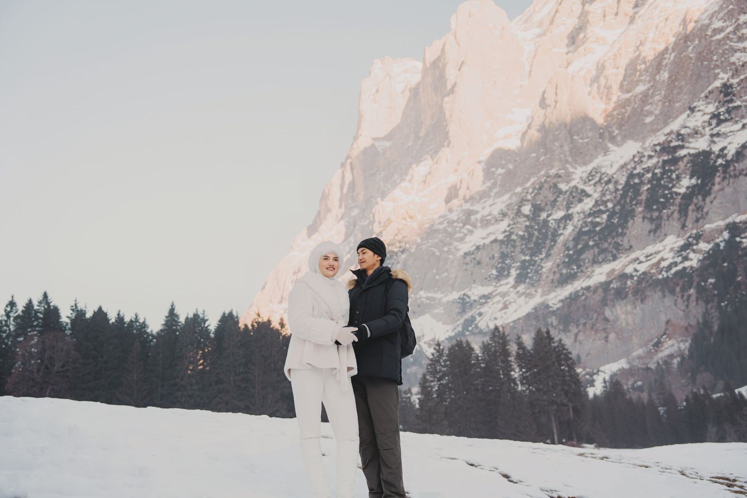 Couple embracing in a vast snowy alpine meadow with towering rocky mountains behind them.