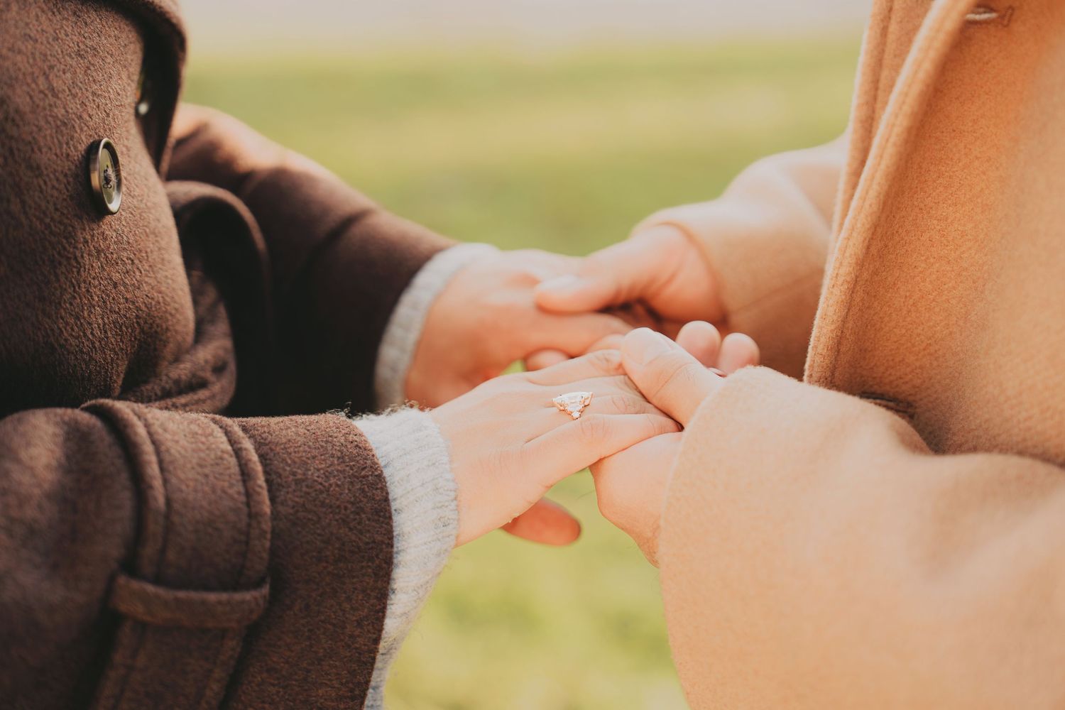 Close-up of couple holding hands outdoors, woman wearing diamond engagement ring, both in warm winter coats.