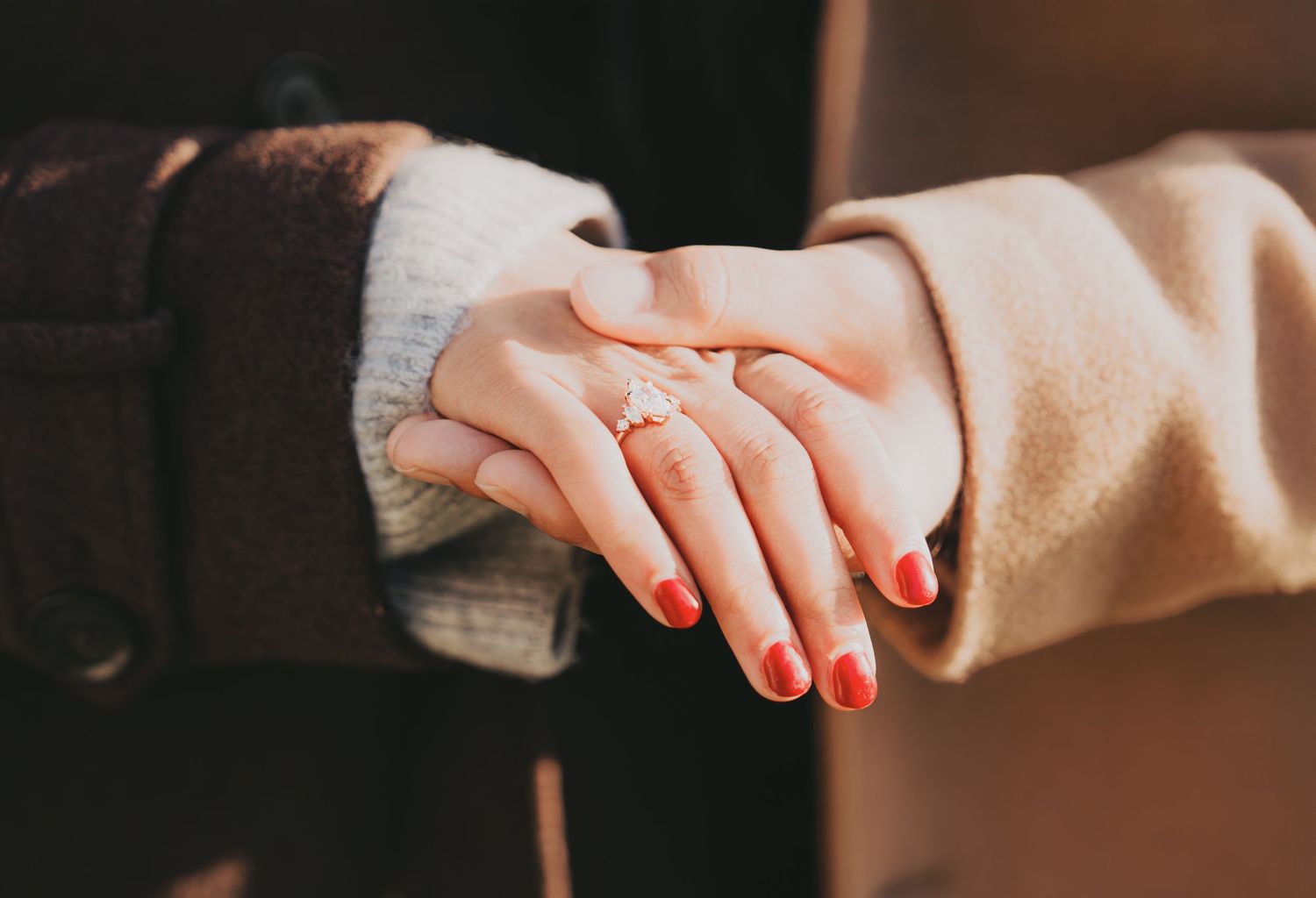 Close-up of woman's hand with red nails and diamond engagement ring held by partner in beige coat.