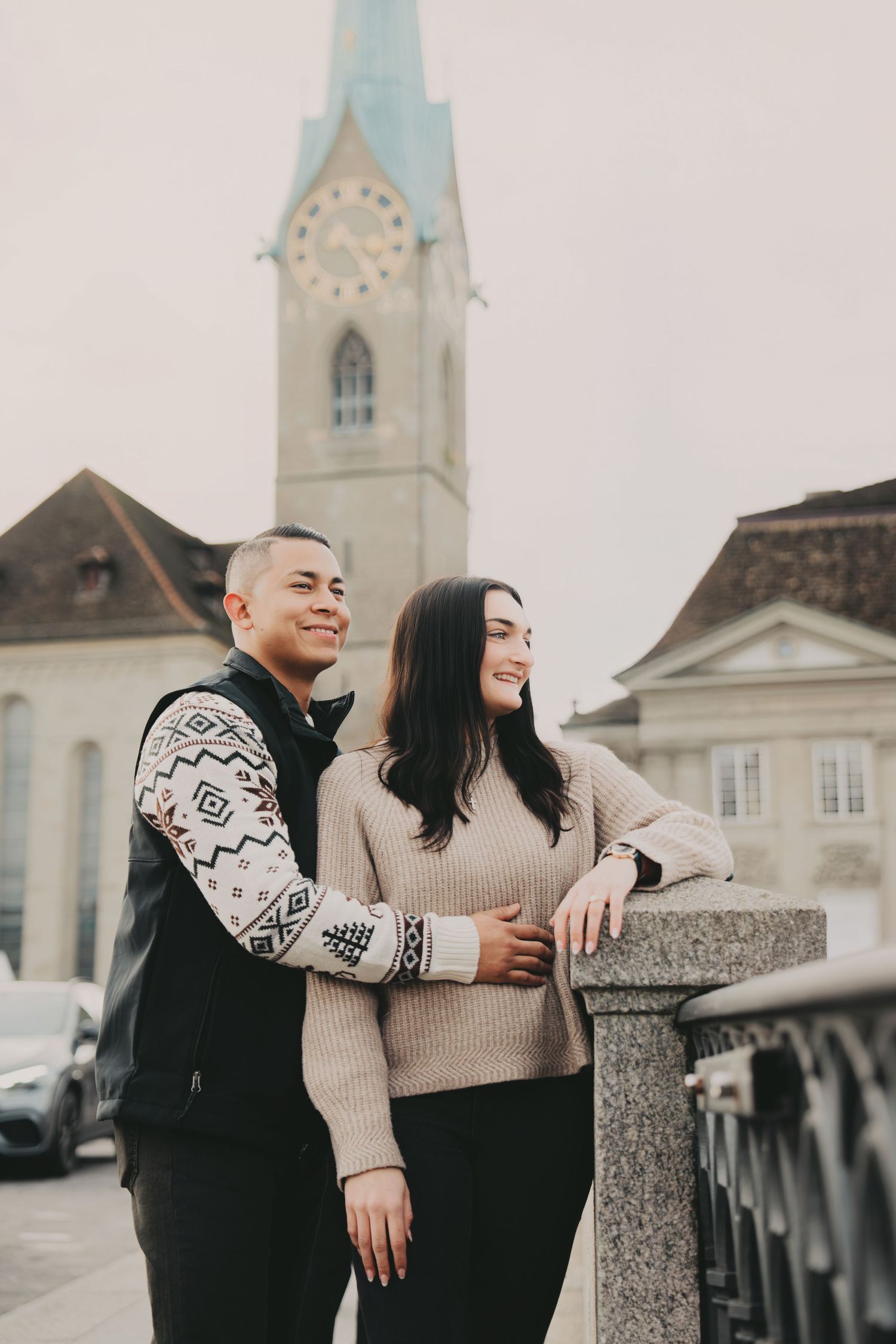Stylish couple standing together on a bridge with Fraumunster Church clock tower visible in Zurich.