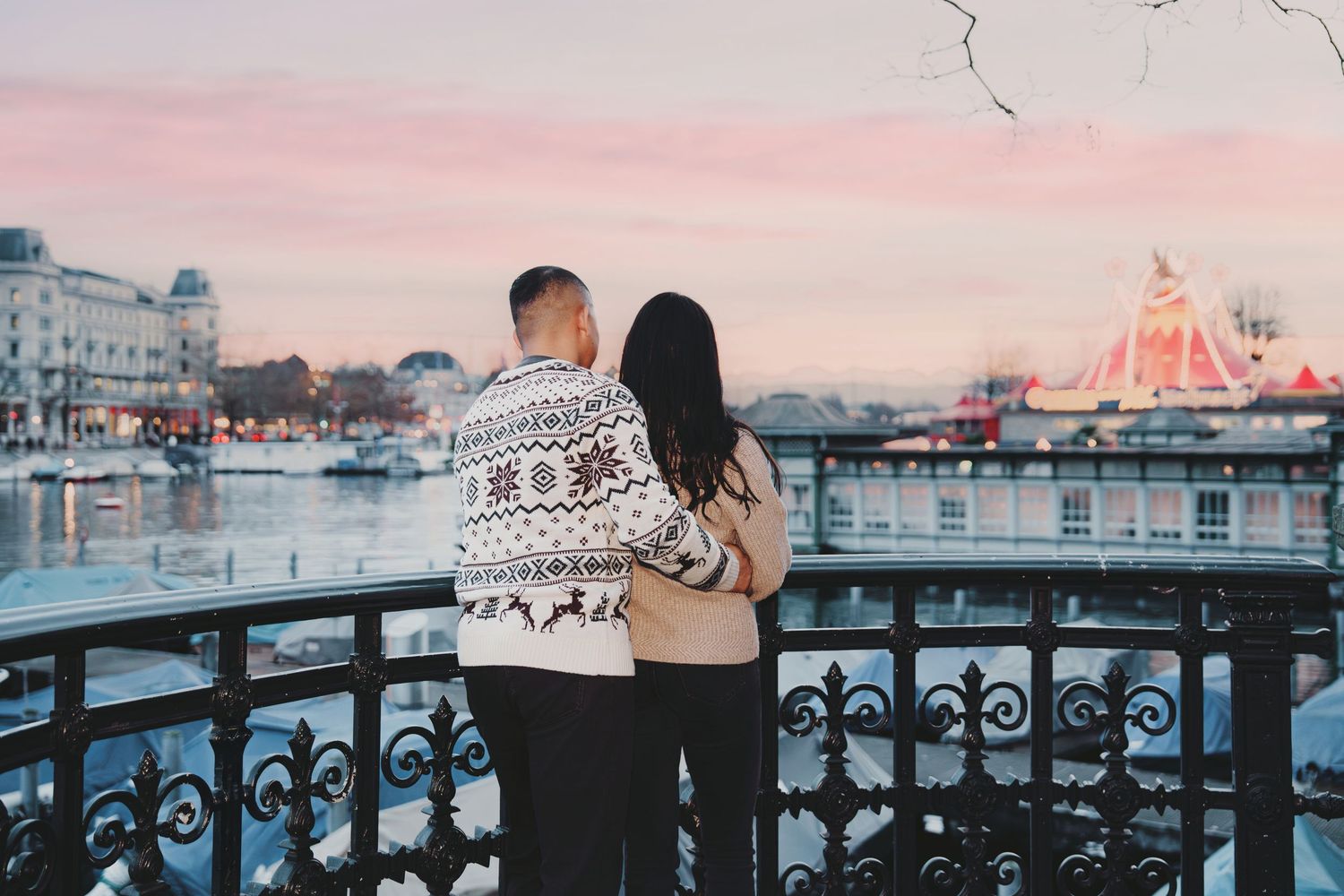 Couple standing arm in arm overlooking Zurich river at sunset with Christmas fair lights.