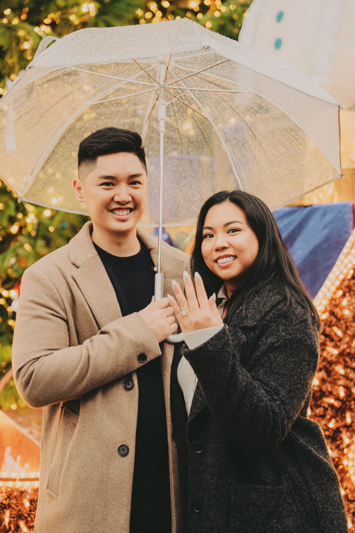 Smiling engaged couple sharing umbrella showing engagement ring outdoors.