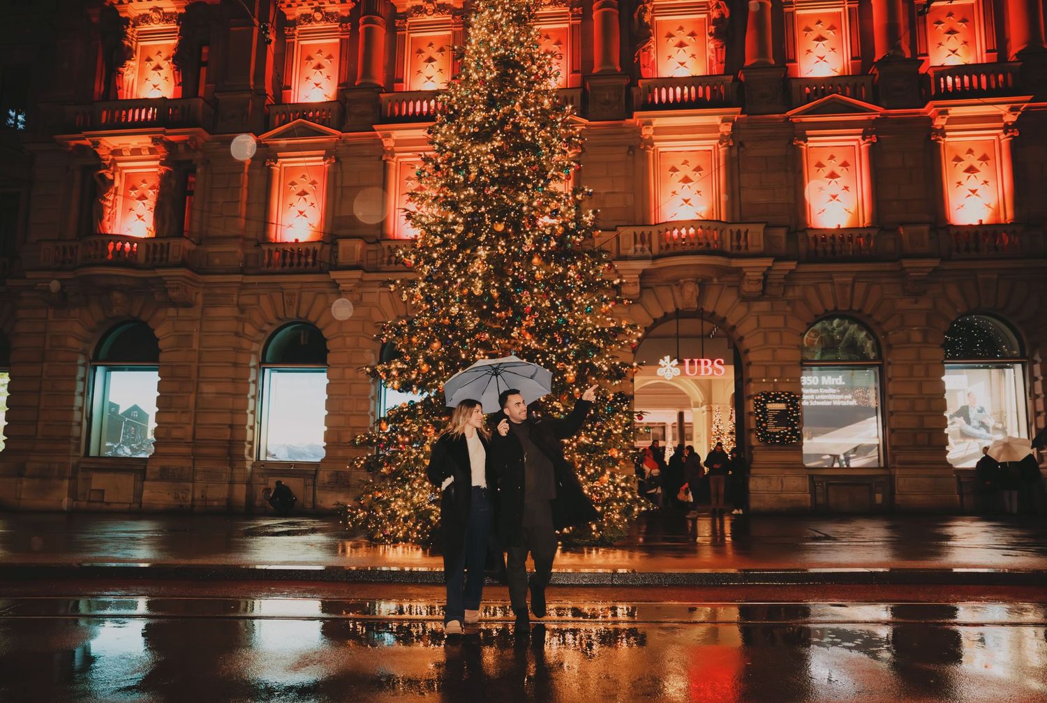 Couple sharing umbrella near large Christmas tree outside illuminated building in rain.