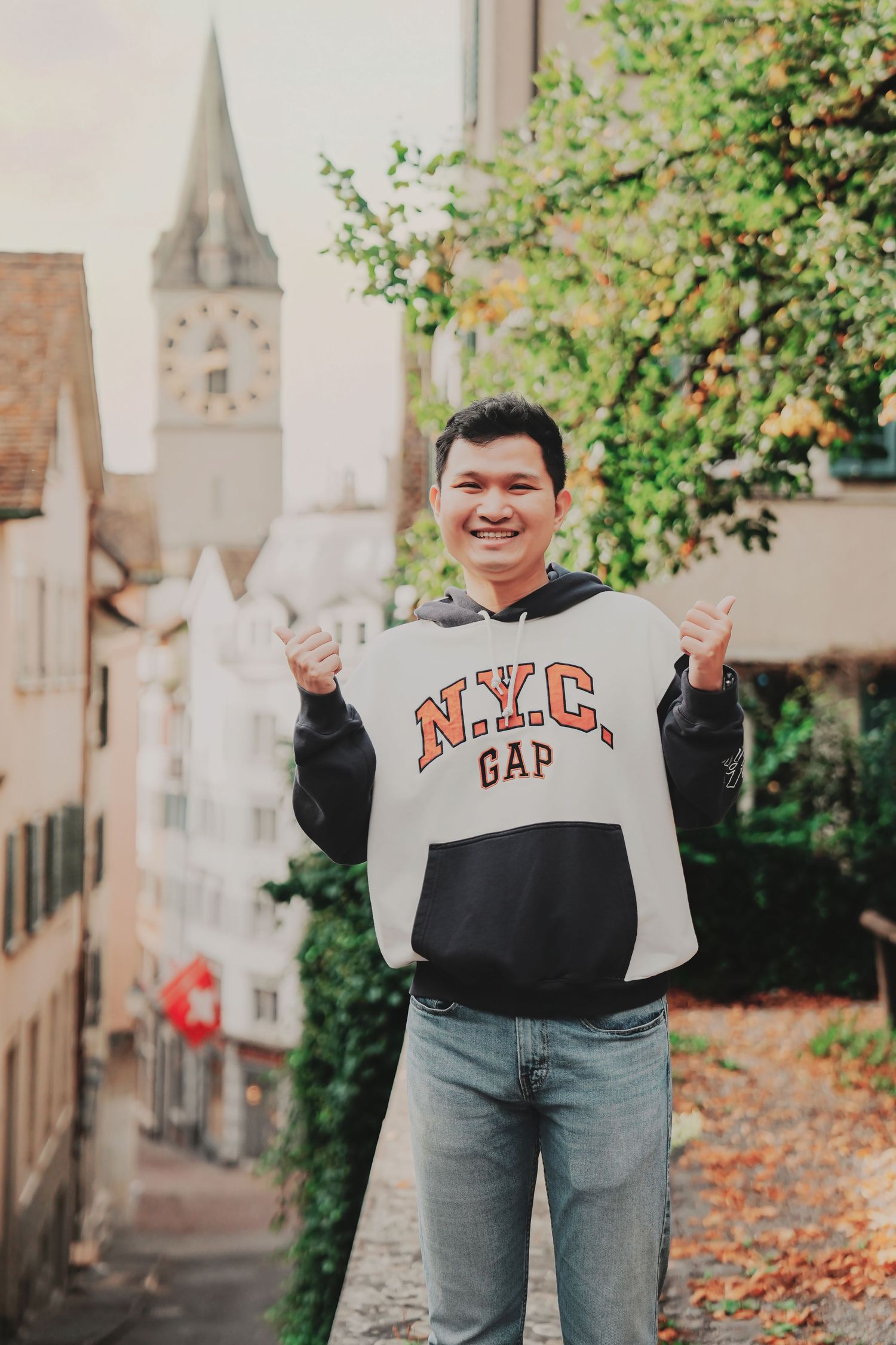 Cheerful man in NYC GAP sweatshirt giving thumbs up near Zurich church tower in autumn.