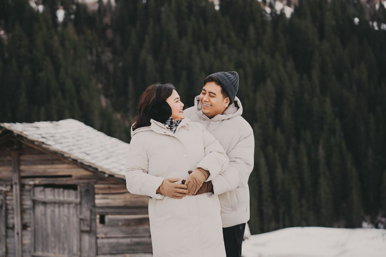 Couple embracing and smiling in winter coats with snow-covered wooden cabin in background.