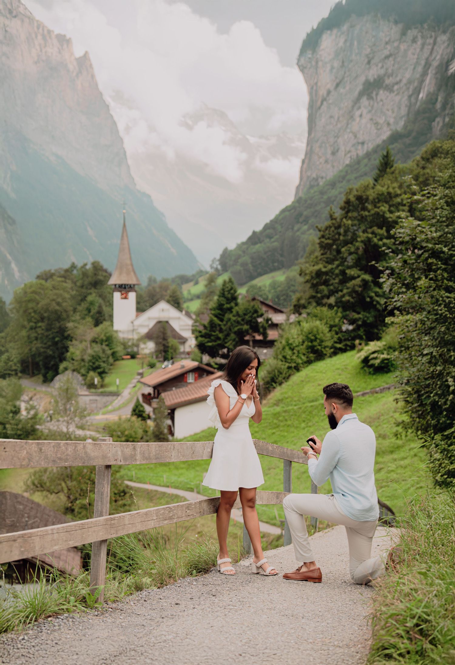 A man proposes on one knee to a surprised woman on a path with a Swiss village and mountains behind them.