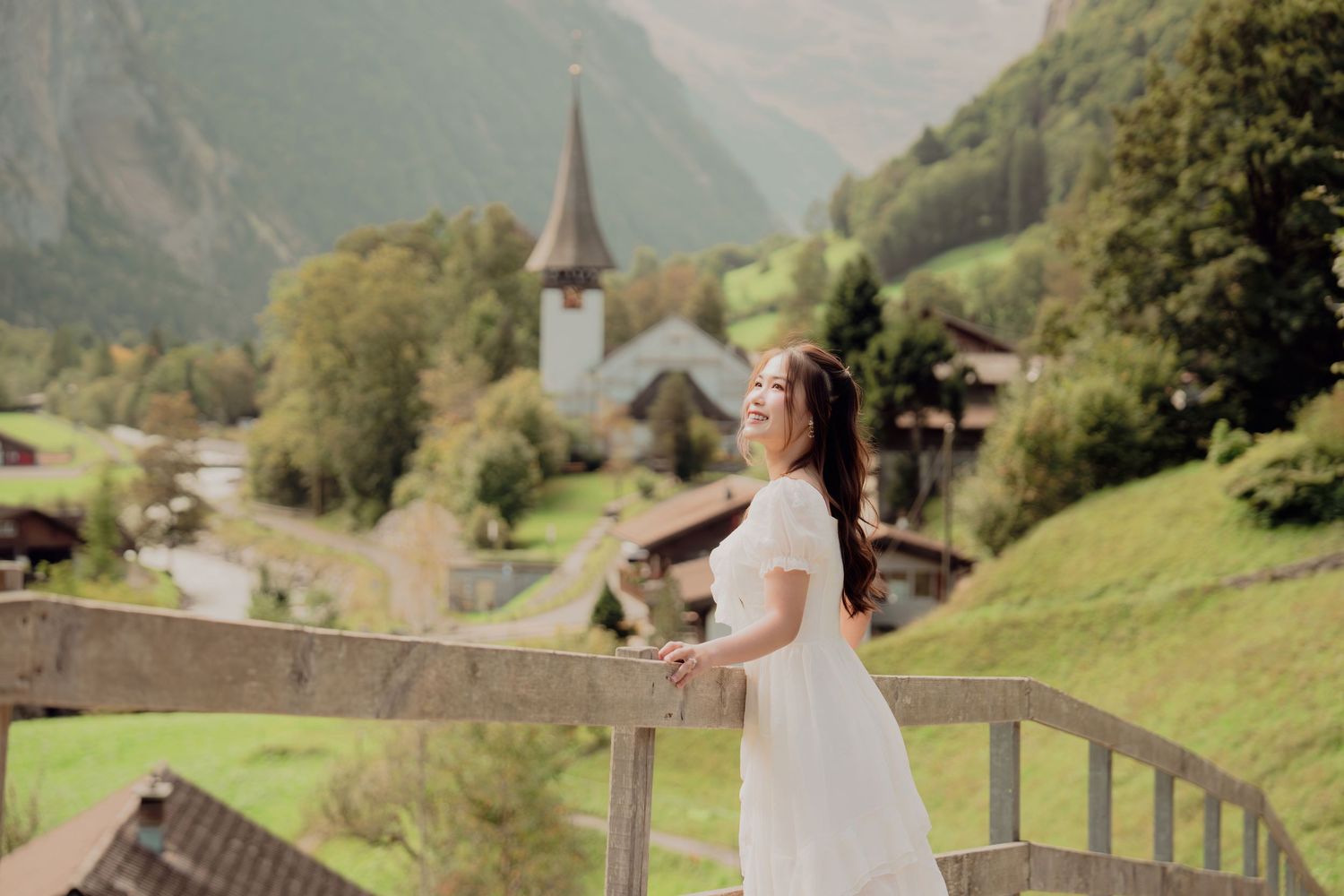Smiling woman in white dress leaning on wooden bridge railing with alpine village and mountains behind.