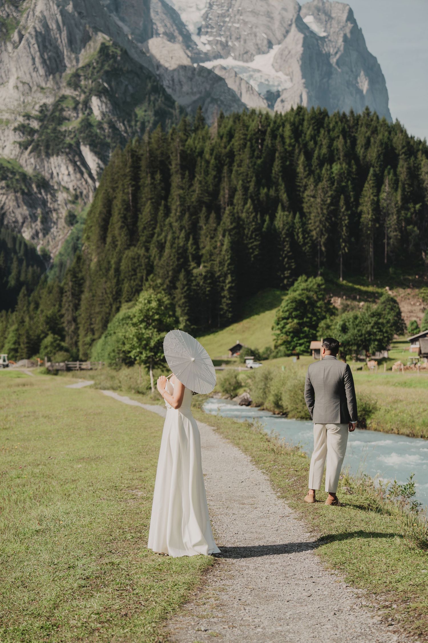 Wedding couple walks along alpine river path surrounded by green meadows and dramatic Swiss mountains.
