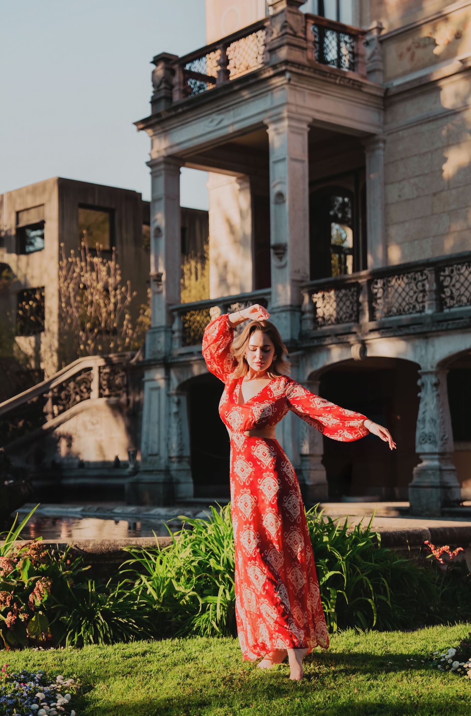 Woman in red patterned maxi dress dances gracefully in garden before an elegant historic building.