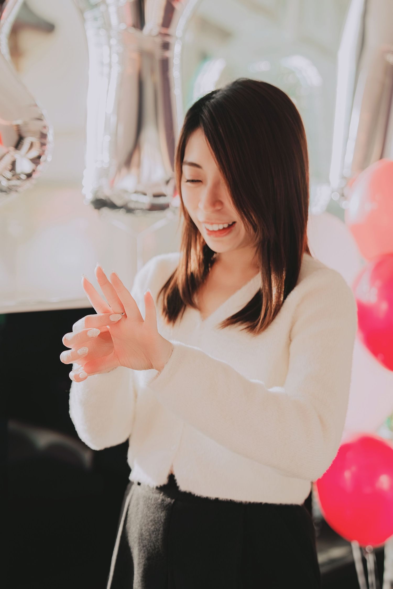 Smiling Asian woman in white sweater admiring engagement ring with pink and silver balloons in background.
