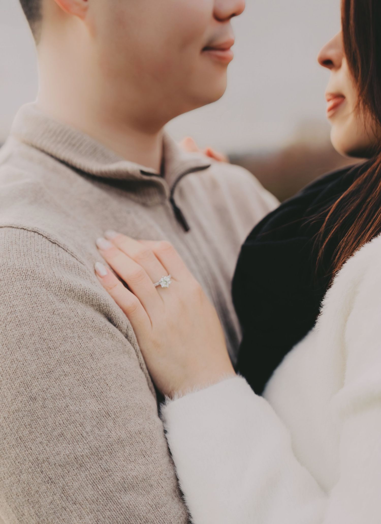 Close-up of womans hand with diamond engagement ring resting on mans beige sweater chest.