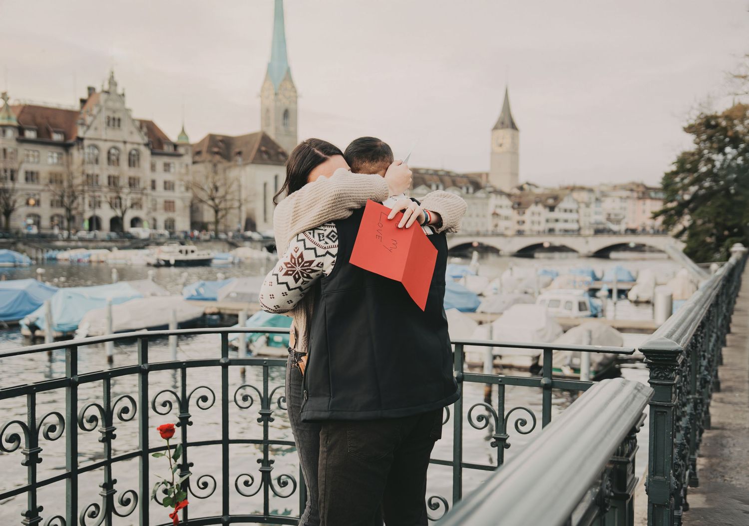 Couple hugging on Zurich bridge holding red gift box with romantic cityscape behind them.