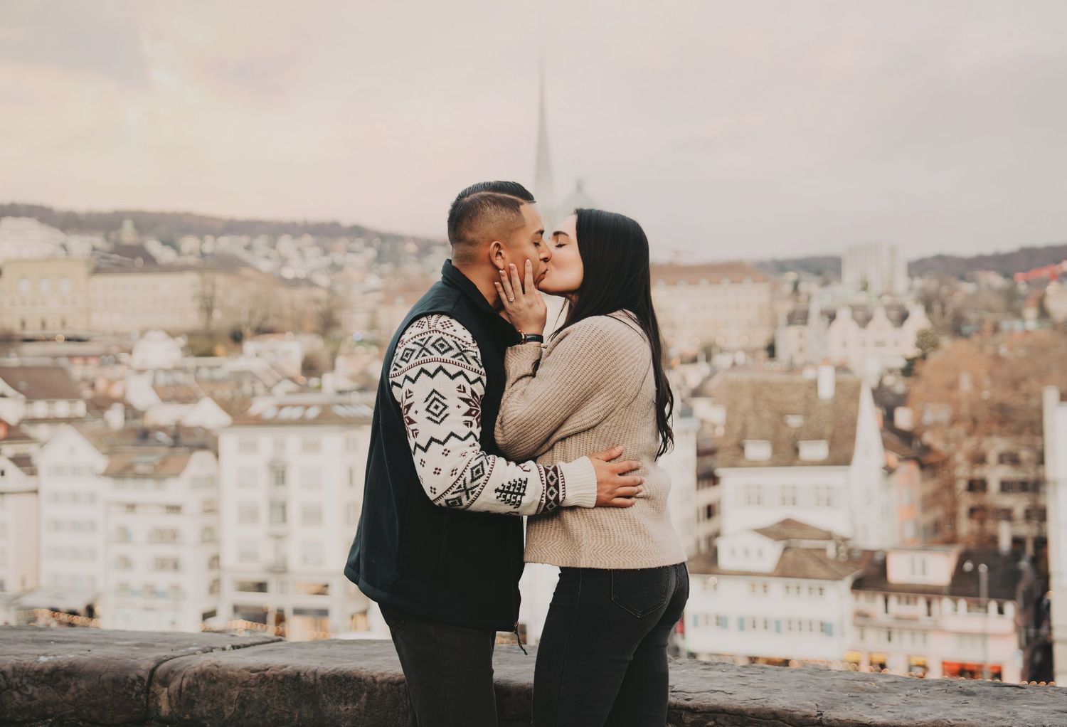 Couple sharing a tender kiss on a stone wall overlooking a European city skyline.