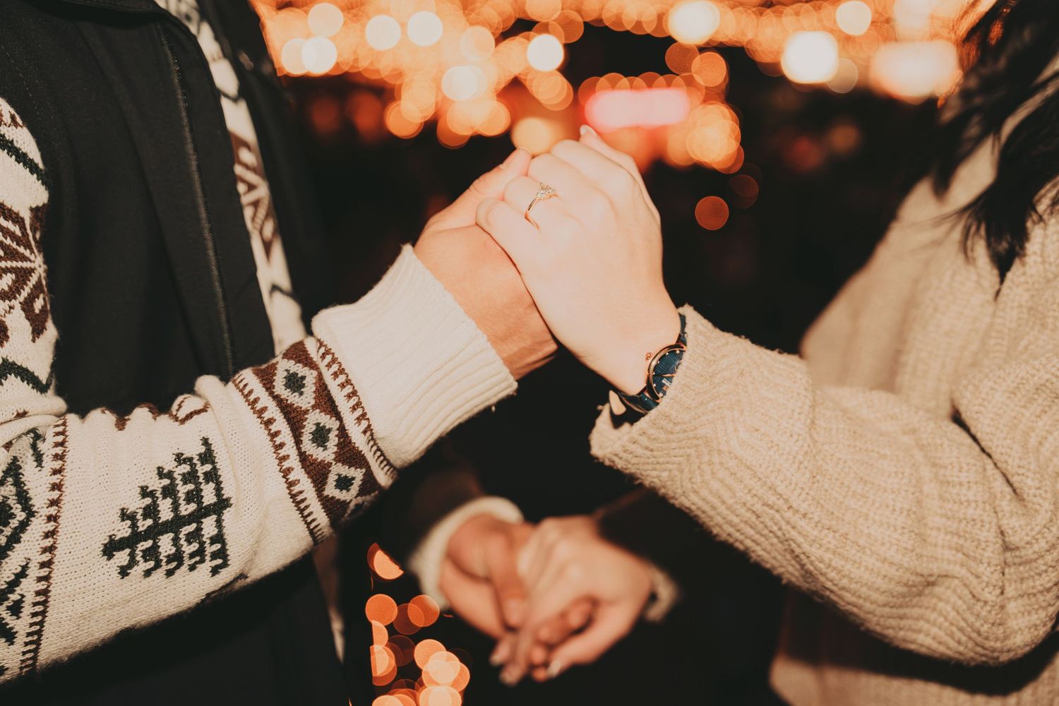 Close-up of couple holding hands wearing cozy winter sweaters with warm bokeh holiday lights behind.