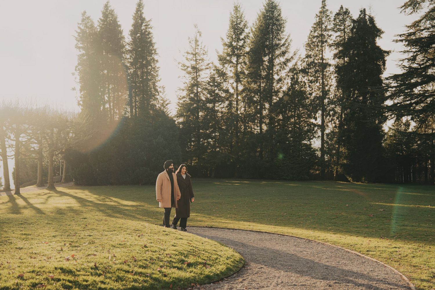 Couple walking hand in hand along a sunlit park path surrounded by tall evergreen trees.