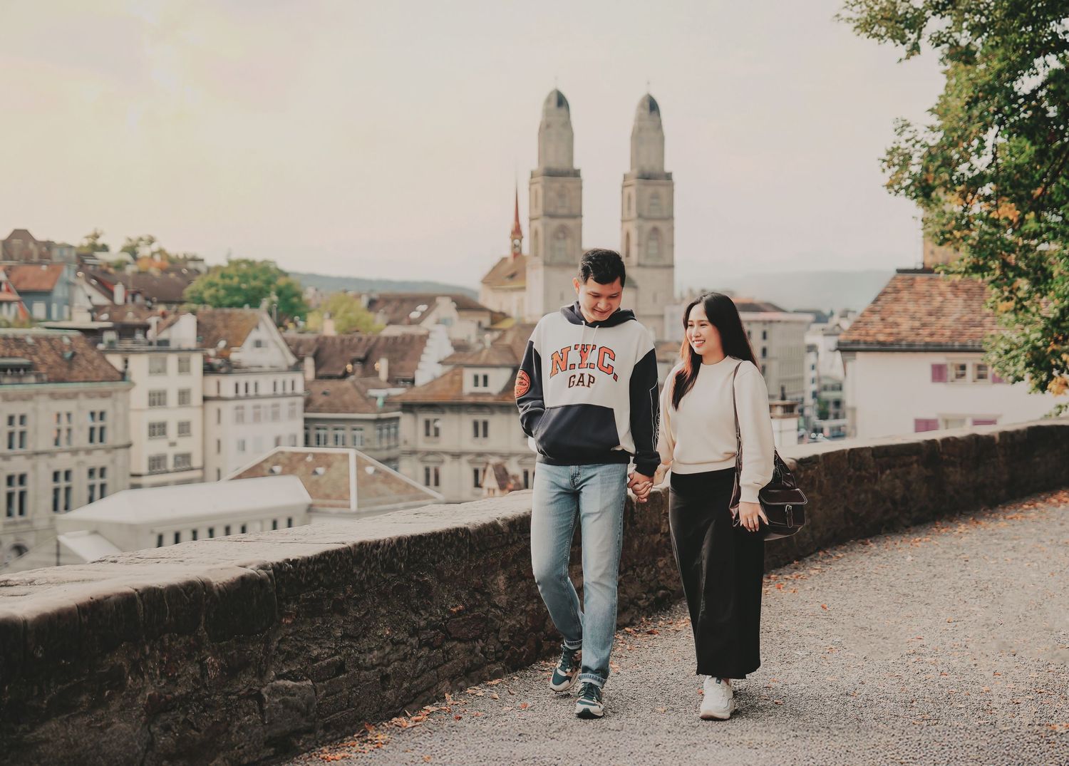 A couple strolls hand-in-hand along a stone wall promenade with Zurich's Grossmünster cathedral in the background.