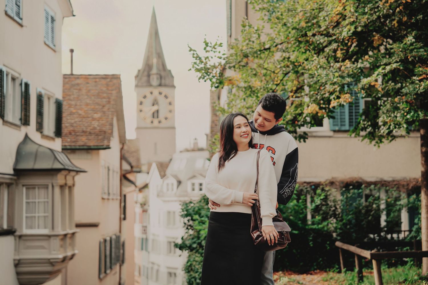 Romantic couple posing together on a scenic overlook in Zurich's old town with St. Peter's Church tower visible.