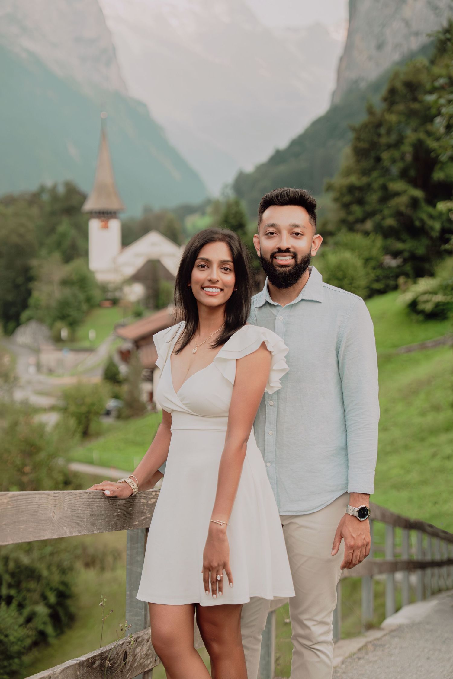 Smiling South Asian couple posing by wooden fence with Swiss alpine village church steeple and mountains in background.