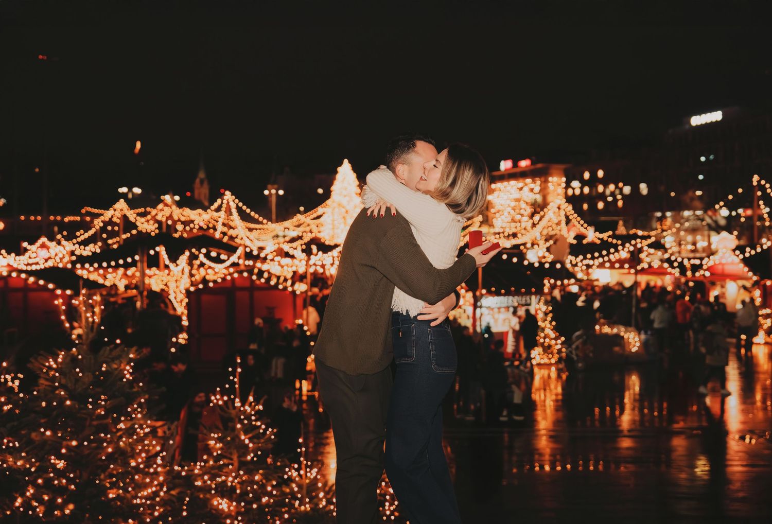 Couple embracing at night in front of beautifully lit Christmas market stalls.