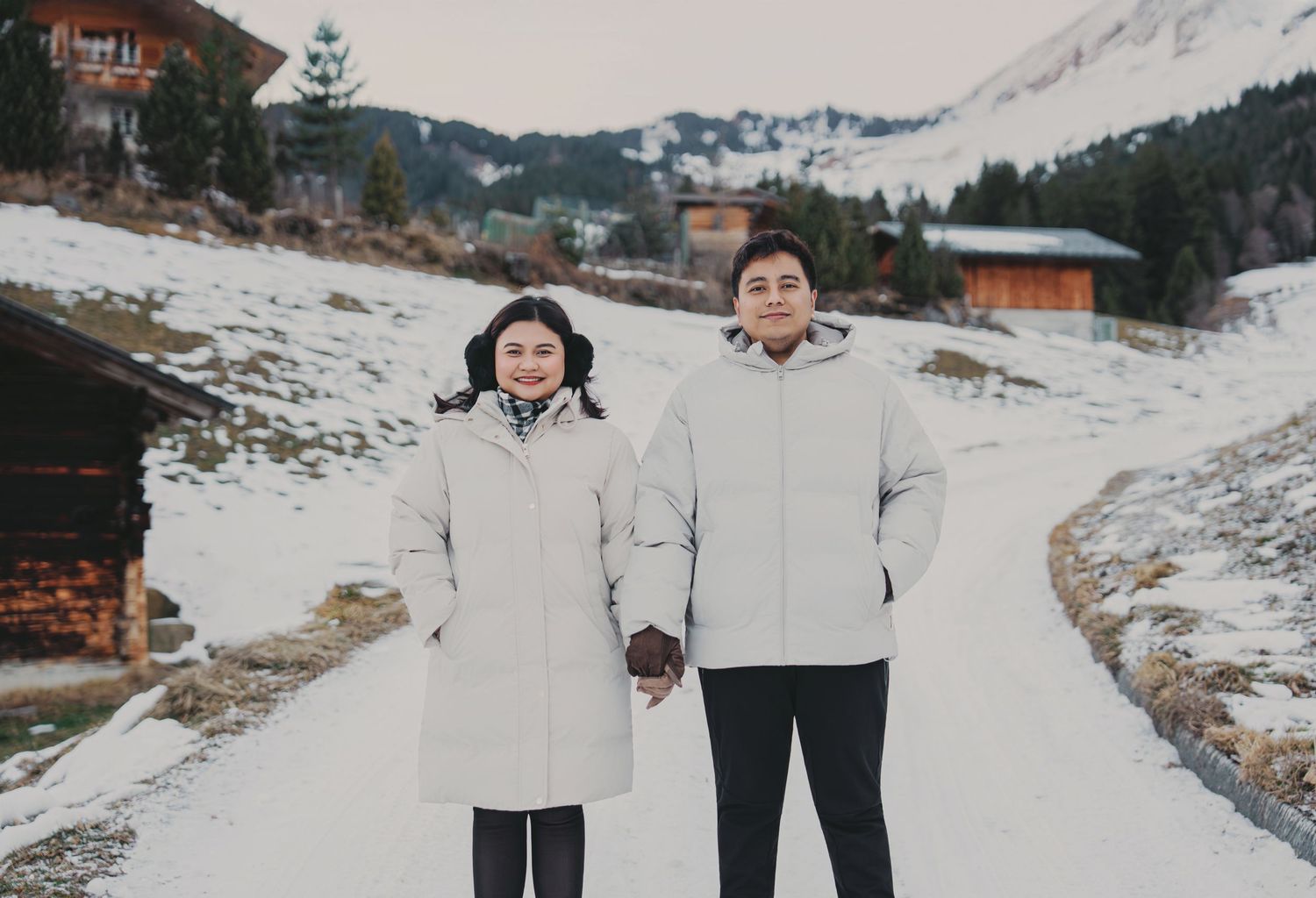 Smiling couple in matching white winter coats holding hands on snowy mountain road.