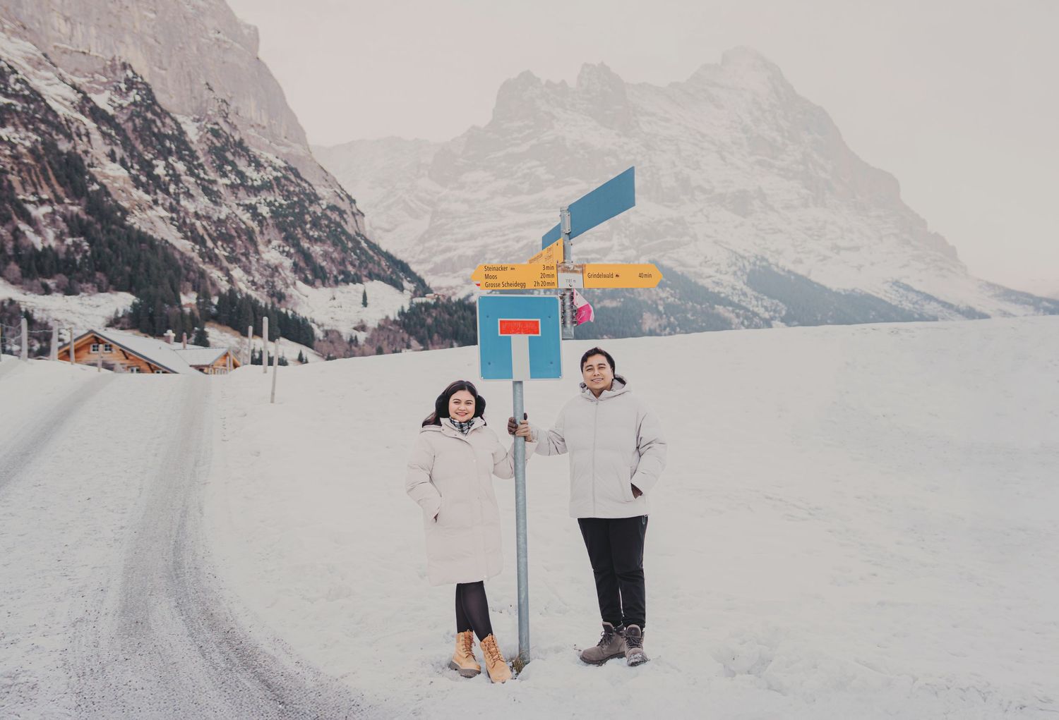 Couple posing beside colorful directional signpost on snowy Swiss alpine road in winter.