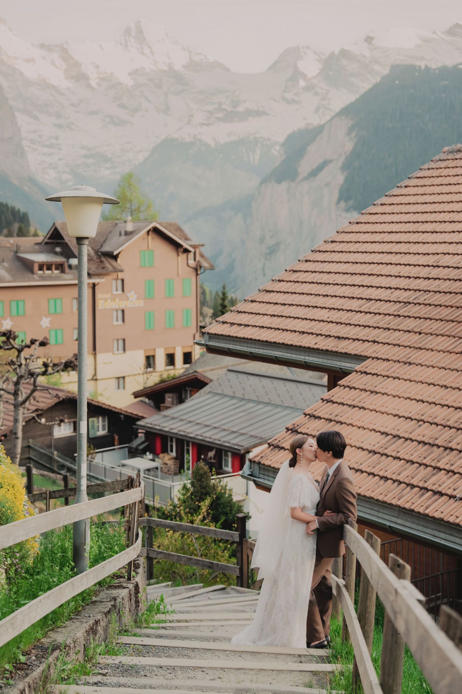Romantic couple sharing a kiss on wooden steps with snow-capped Swiss Alps and village rooftops behind them.