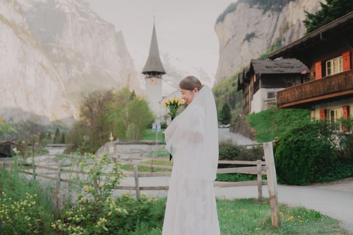 Bride in white wedding gown smells yellow bouquet beside a rustic Swiss alpine church.