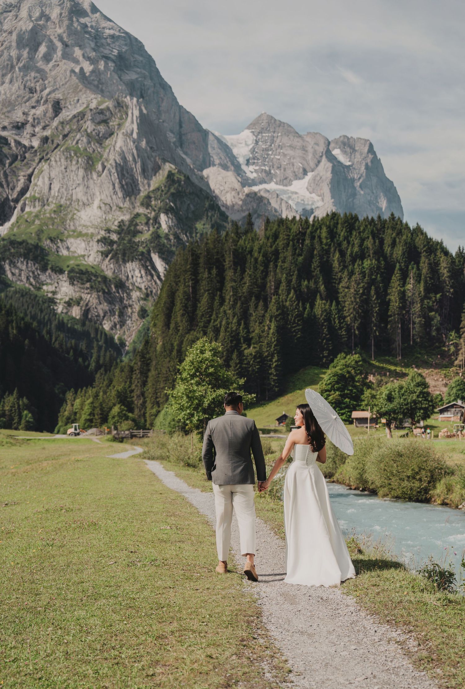 Couple in wedding attire walking hand-in-hand along a mountain path with a white parasol in the Swiss Alps.