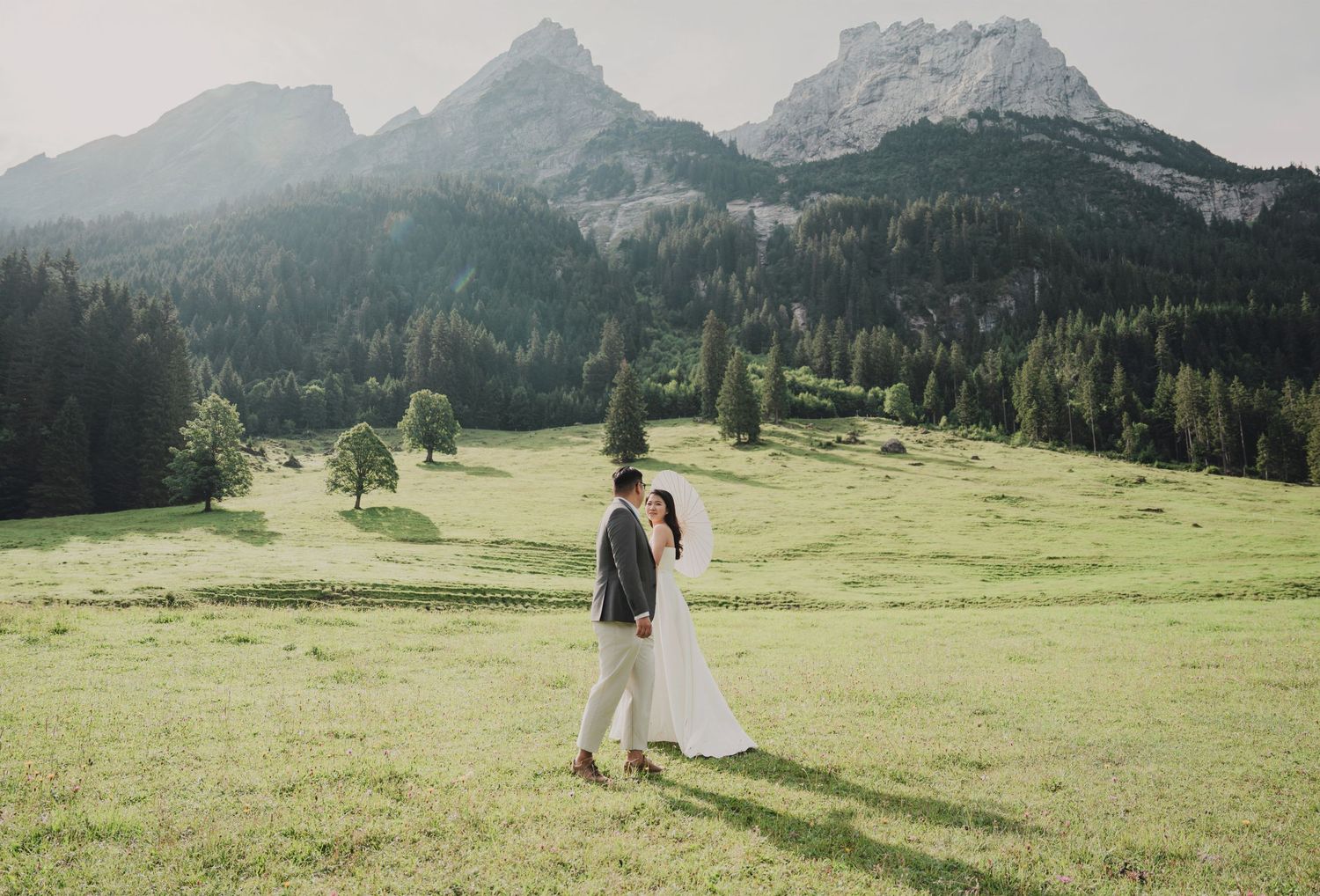 Bride and groom embrace in sunlit alpine meadow surrounded by pine trees and majestic mountain peaks.
