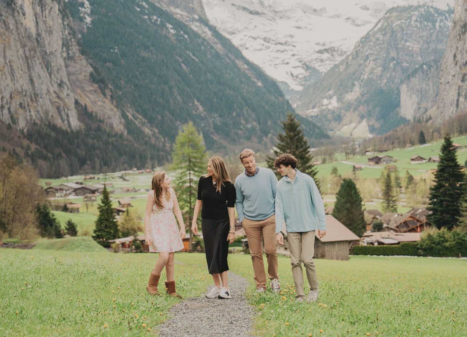 Family of four strolling on a path through a picturesque Swiss mountain valley with snowy peaks behind.
