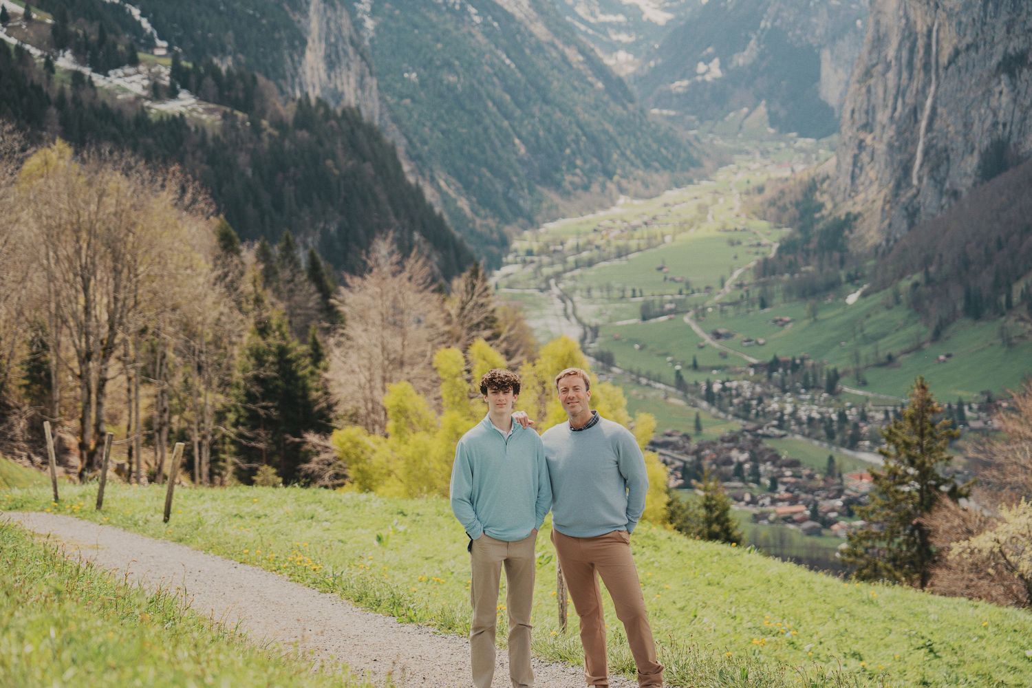 Two young men in matching light blue sweaters stand on hillside path in Swiss Alps valley.