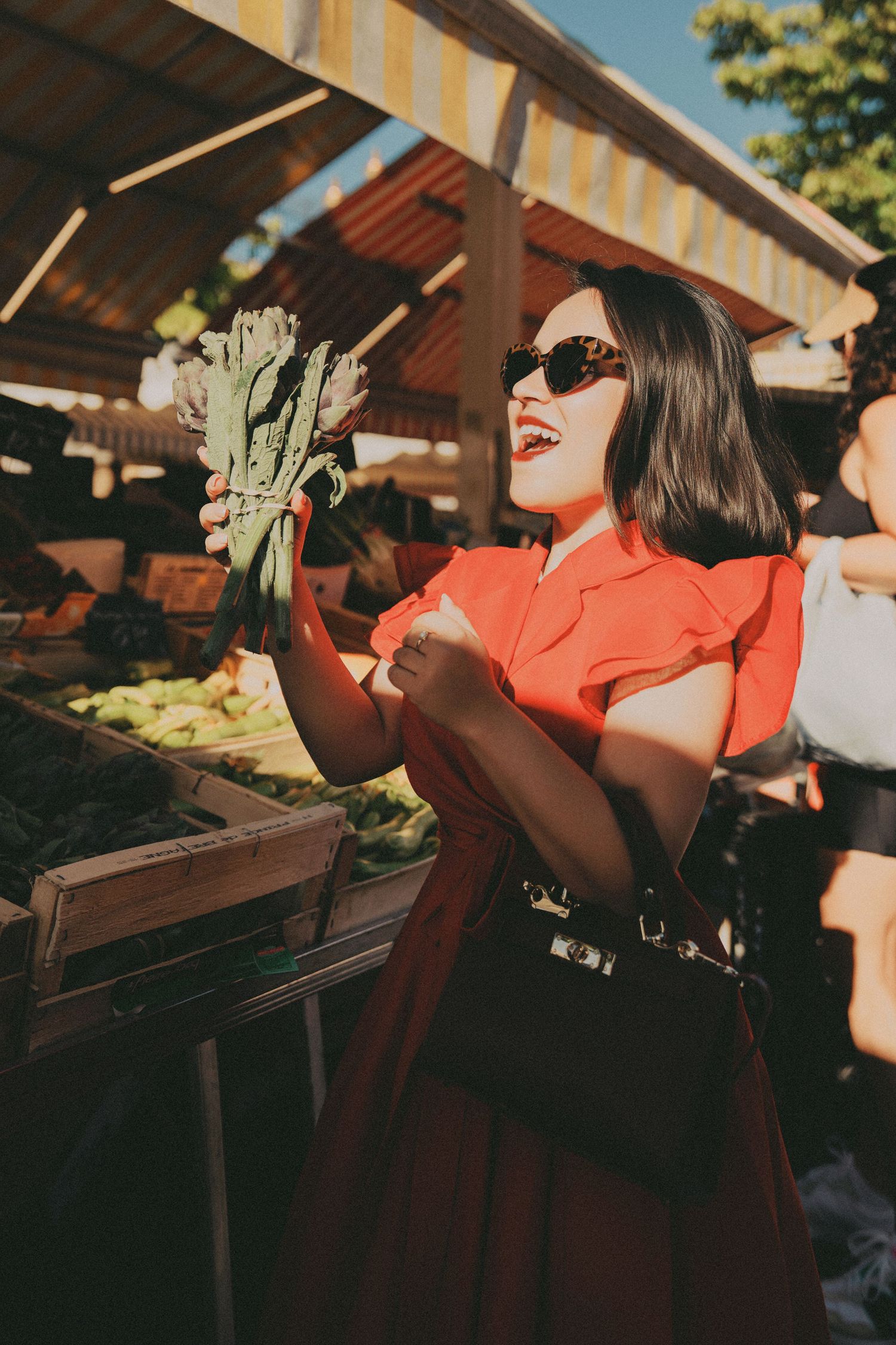 Smiling woman in red dress and sunglasses holding vegetables at sunny outdoor farmers market.