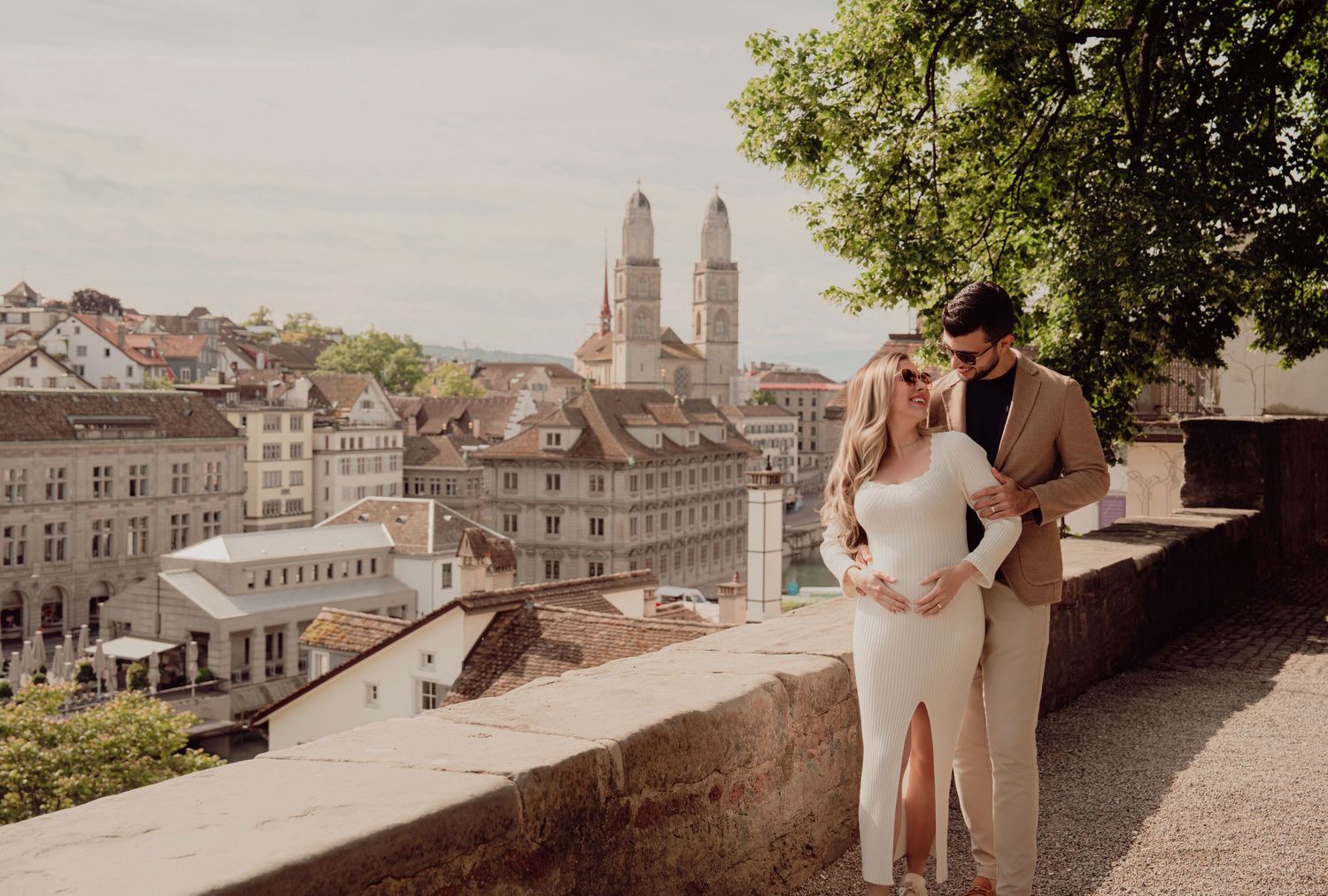 Expecting couple embraces on terrace with Zurich old town skyline and Grossmünster cathedral behind.