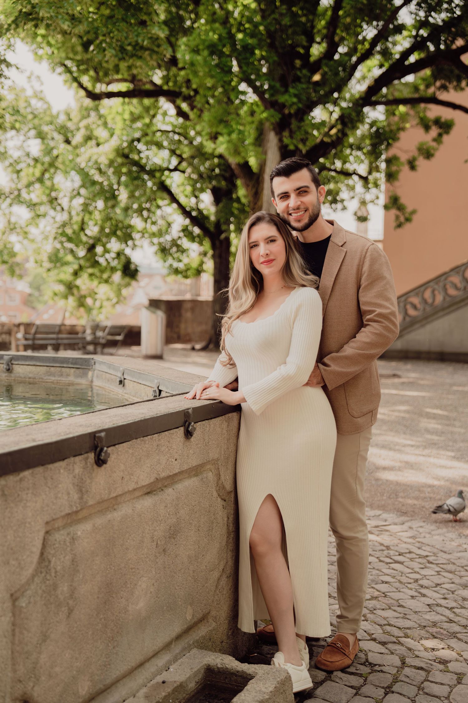 Happy couple posing by a stone fountain, woman in elegant white slit dress, man in tan blazer embracing her.