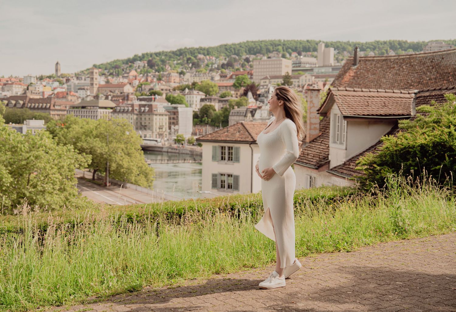 Pregnant woman in white outfit standing on cobblestone path with scenic European city view behind.