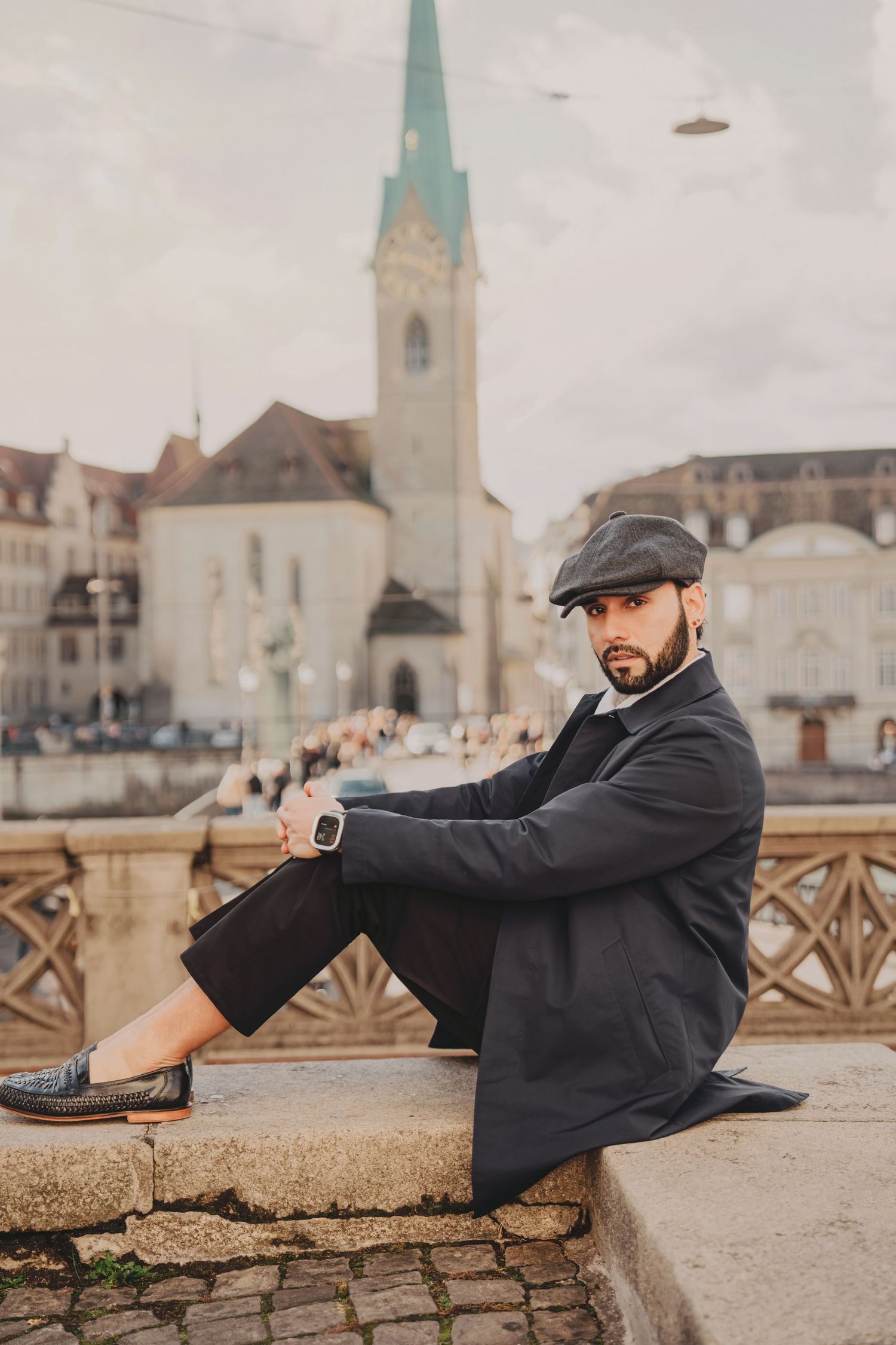 Trendy bearded man in dark coat and flat cap sits on stone steps with Zurich church spire in background.