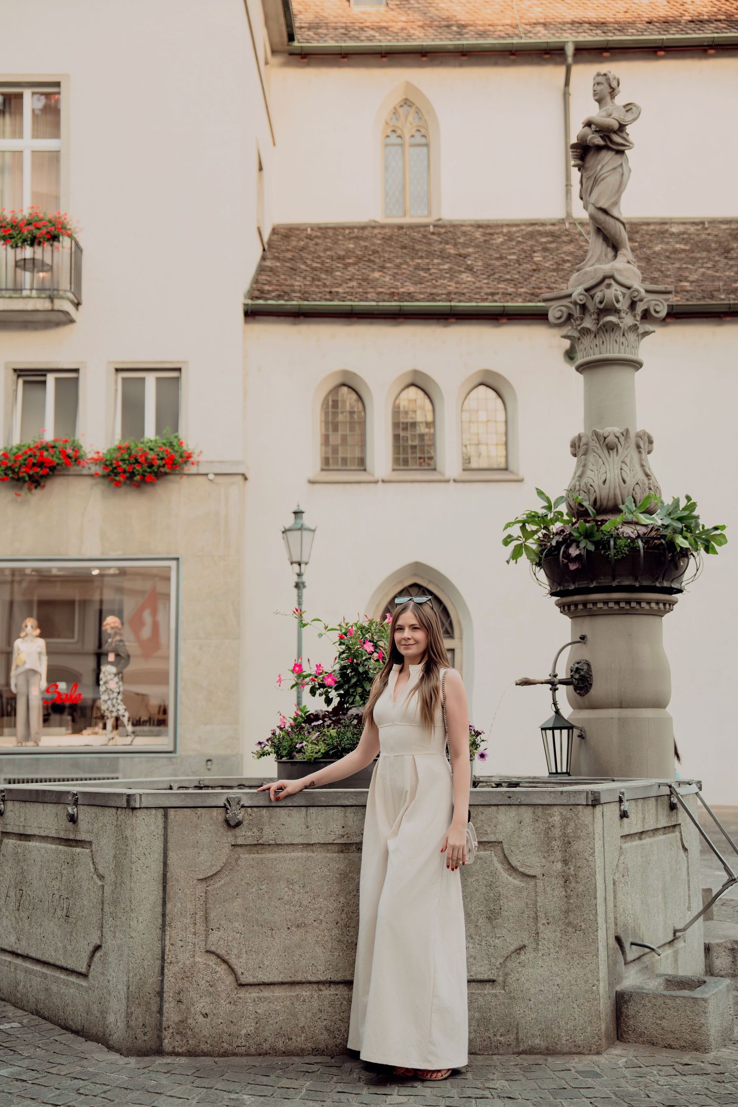 Woman in elegant white maxi dress standing by ornate stone fountain in charming European old town square.
